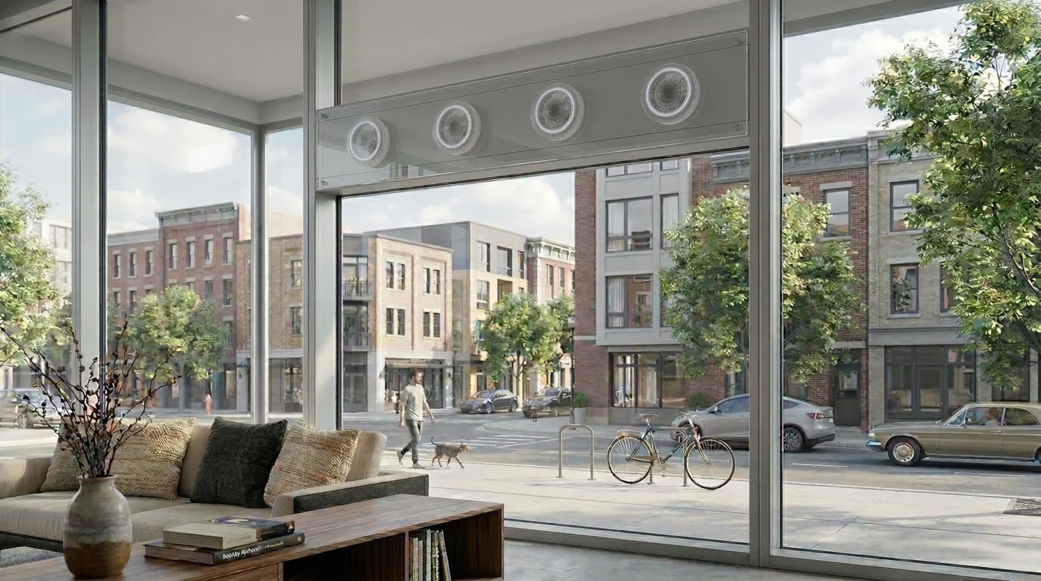 Vista interior de un moderno apartamento urbano iluminado por la luz del día, que muestra una gran ventana de suelo a techo con vistas al tráfico de la ciudad. Un dispositivo rectangular de metal, que representa la tecnología de cancelación activa de ruido (ANC) está montado en el travesaño central de la ventana.